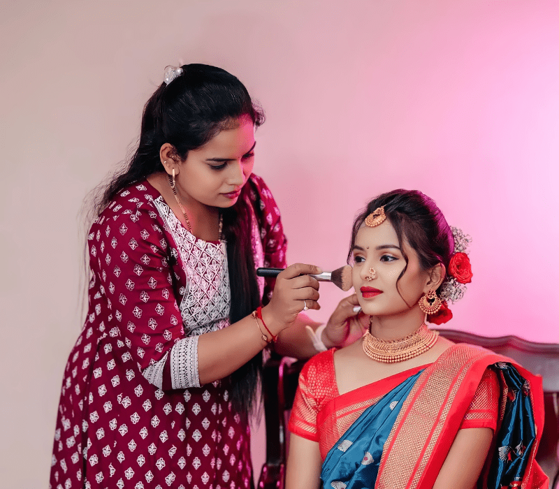 the girl is sitting on the chair and another girl was doing makeup